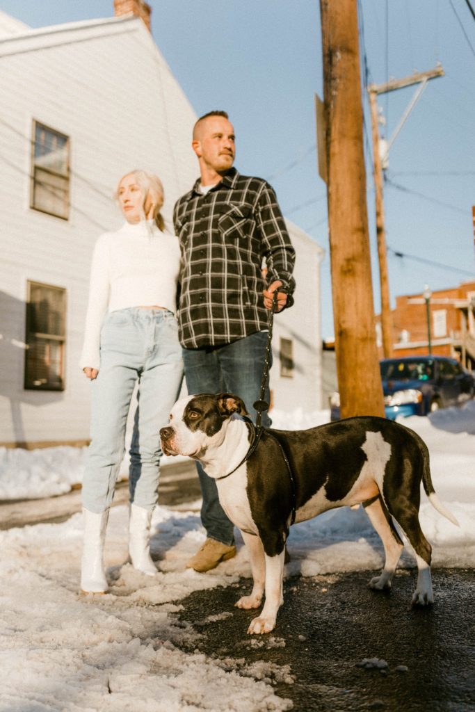 a couple pose with their dog on a snow covered street for engagement session 