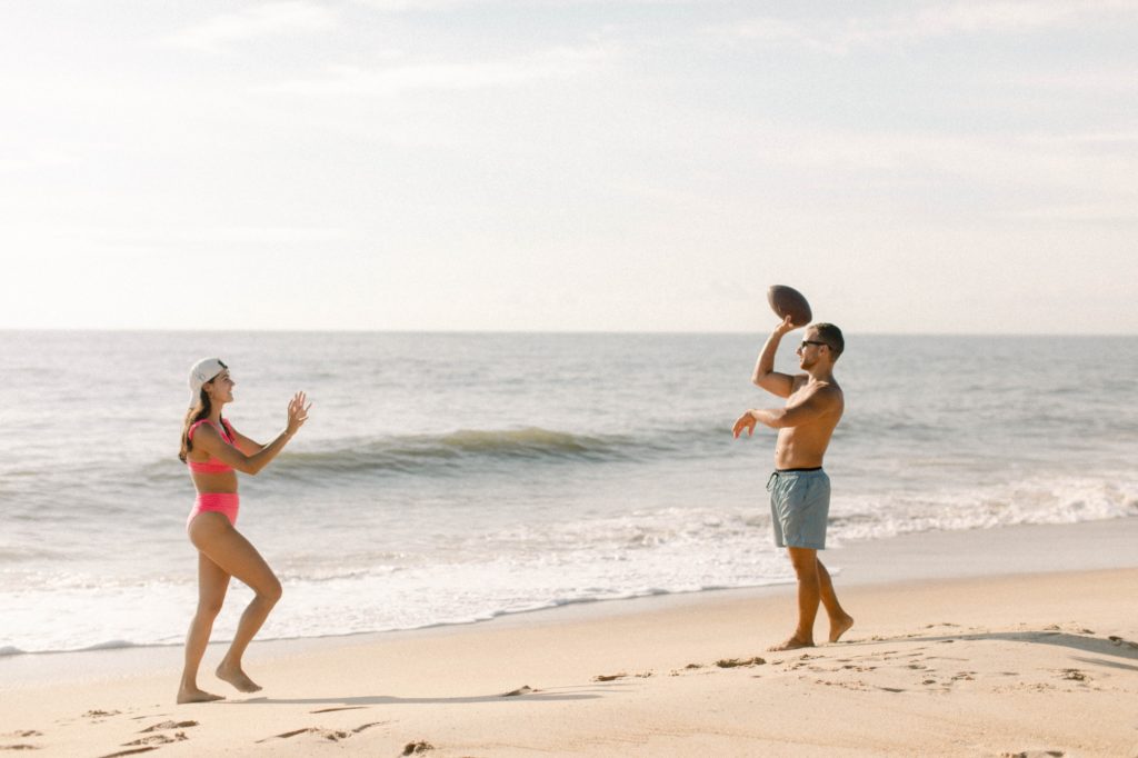 a couple throws football on beach in bathing suits for sunrise rehoboth beach engagement session