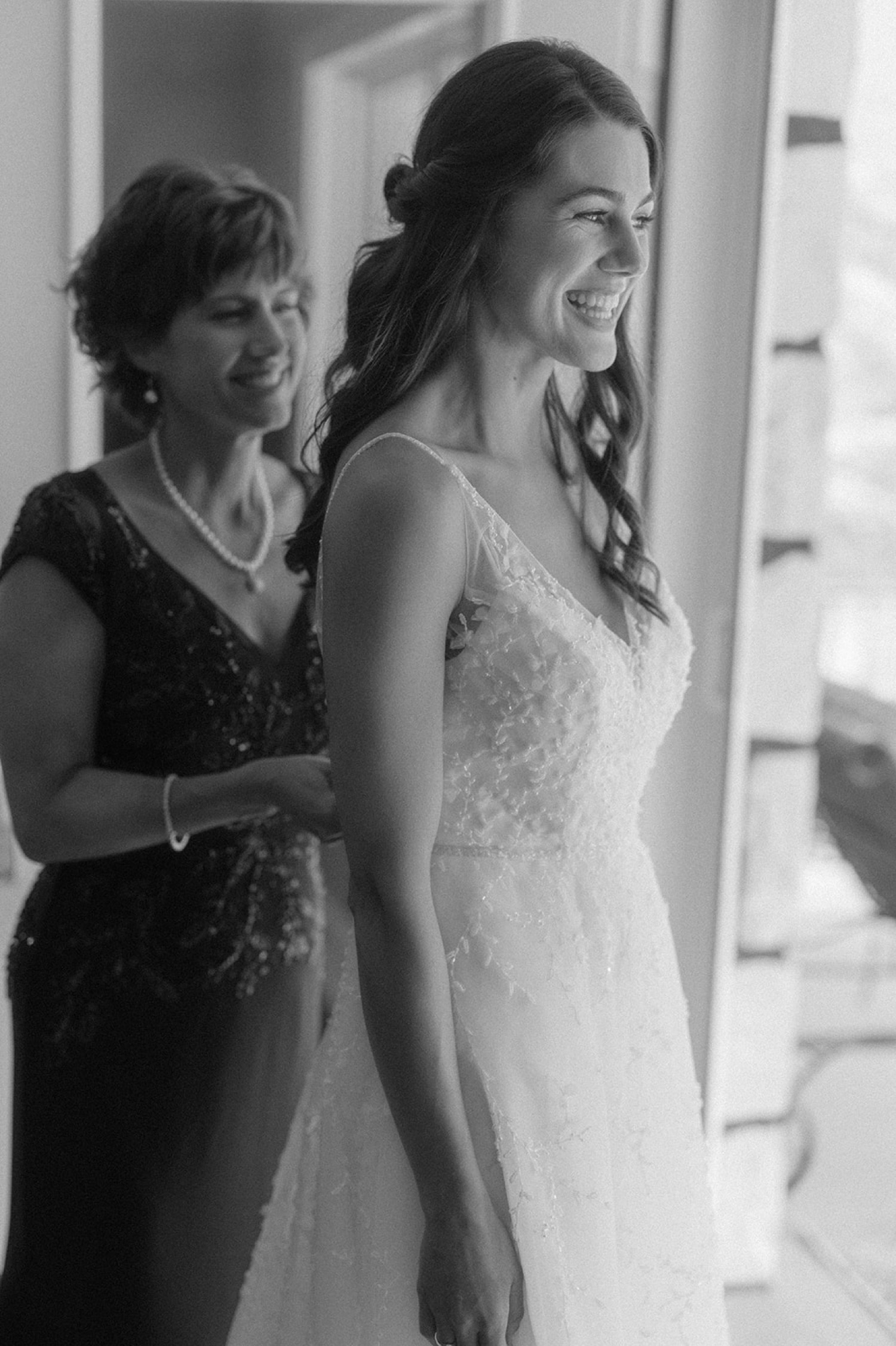 bride and mom smiling buttoning wedding dress