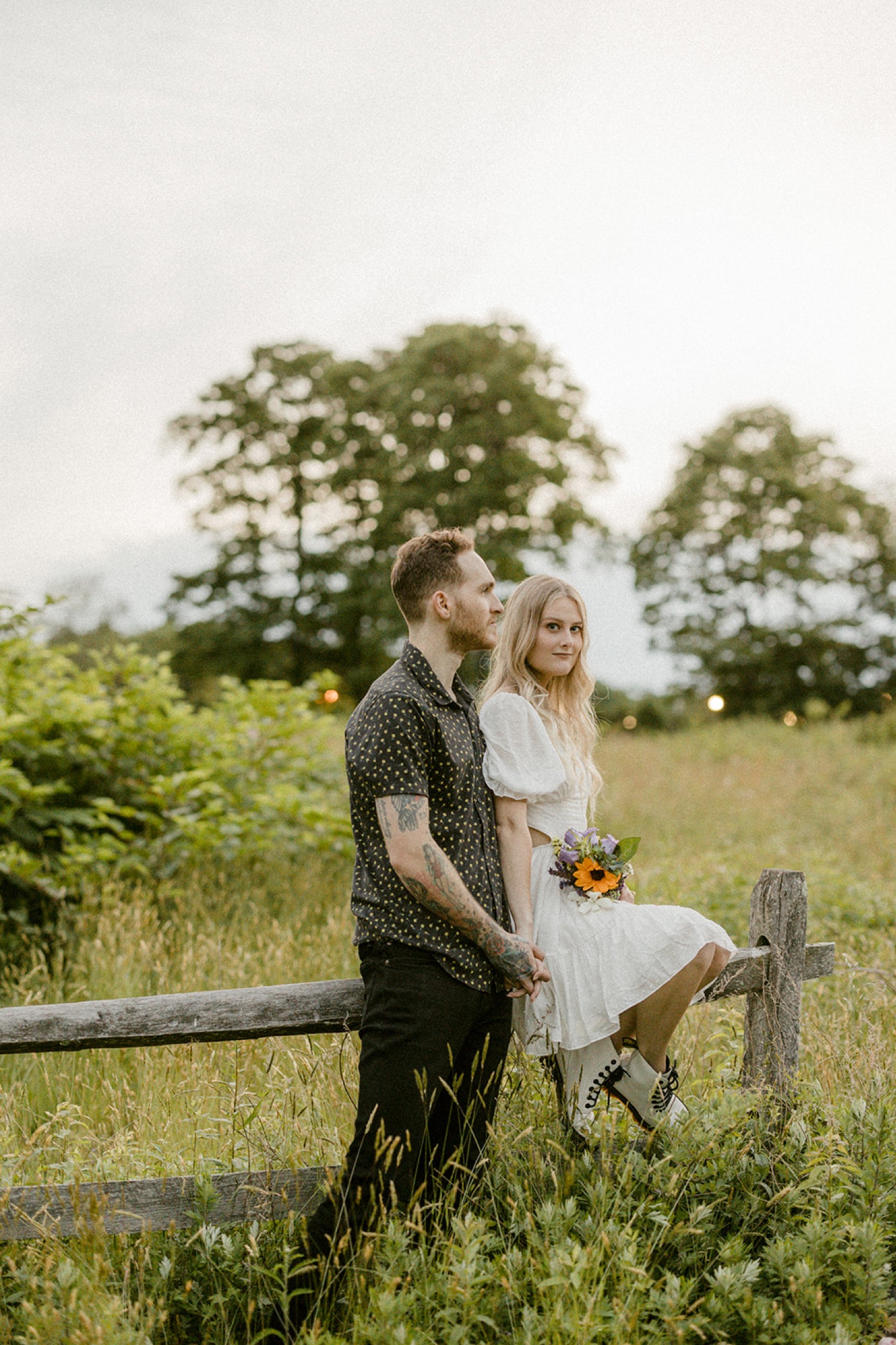 couple sitting on fence in field holding flowers boho chic engagement session andorra meadows